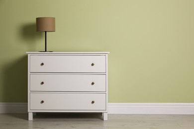 Modern white chest of drawers with lamp near light green wall indoors. Space for text