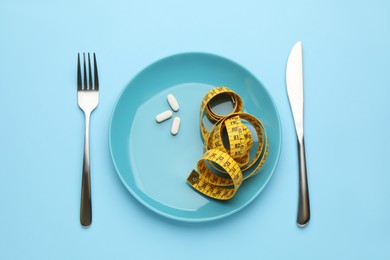 Plate with weight loss pills, measuring tape and cutlery on light blue background, flat lay