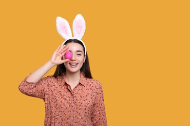 Photo of Happy woman in bunny ears headband holding painted Easter egg on orange background. Space for text