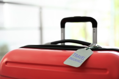 Stylish suitcase with travel insurance label on blurred background, closeup