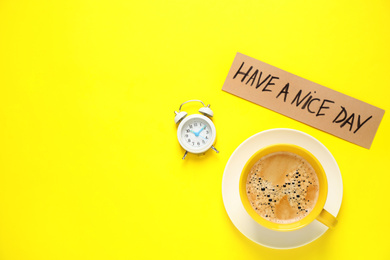 Delicious morning coffee, alarm clock and card with HAVE A NICE DAY wish on yellow background, flat lay. Space for text
