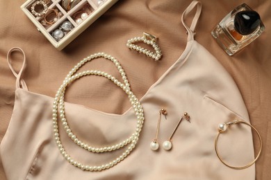 Stylish jewelry with pearls, luxury perfume and silk dress on beige fabric, flat lay