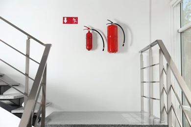 Fire extinguishers and emergency exit sign on white wall near staircase indoors
