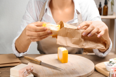 Woman putting natural handmade soap into paper bag at wooden table, closeup