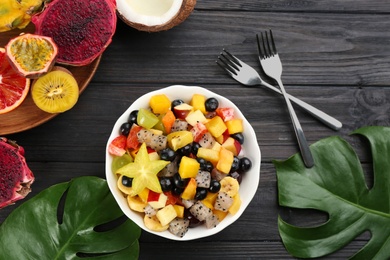 Delicious exotic fruit salad and ingredients on black wooden table, flat lay