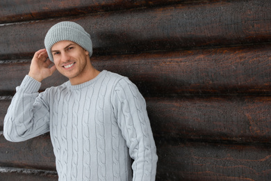 Handsome man wearing warm sweater and hat near wooden wall, space for text. Winter season