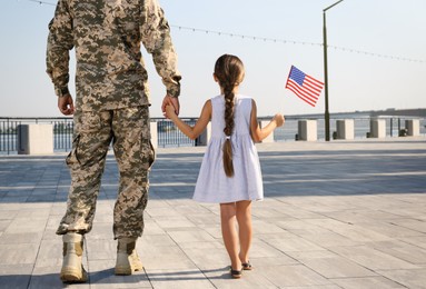 Soldier and his little daughter with American flag outdoors, back view. Veterans Day in USA
