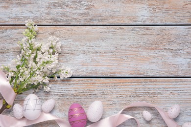 Photo of Many painted Easter eggs, branch of lilac flowers and ribbon on rustic wooden table, flat lay. Space for text