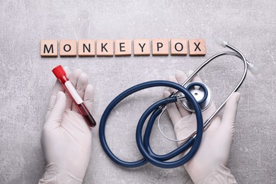 Word Monkeypox made of wooden squares, top view. Doctor holding test tube and stethoscope at light grey table, closeup
