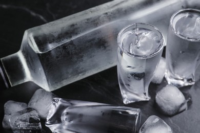 Bottle of vodka and shot glasses with ice on black table, closeup