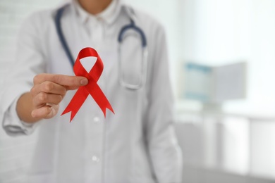 Photo of Doctor holding red ribbon on blurred background, closeup with space for text. AIDS awareness month