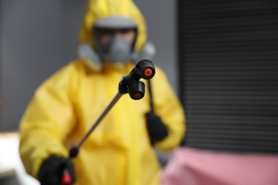 Pest control worker in protective suit indoors, focus on insecticide sprayer