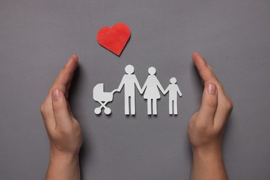 Man protecting paper family figures and red heart on grey background, top view. Insurance concept