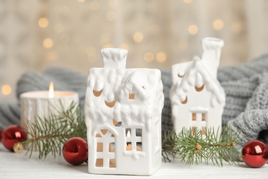 House shaped holders with burning candles on white wooden table against blurred Christmas lights