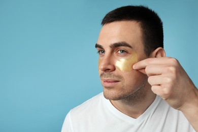 Young man applying under eye patches on light blue background. Space for text