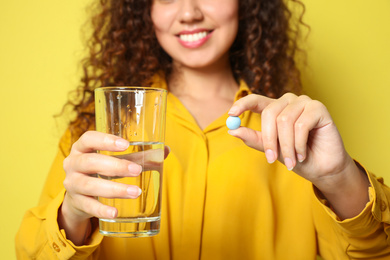 African-American woman with glass of water and vitamin pill against yellow background, focus on hands