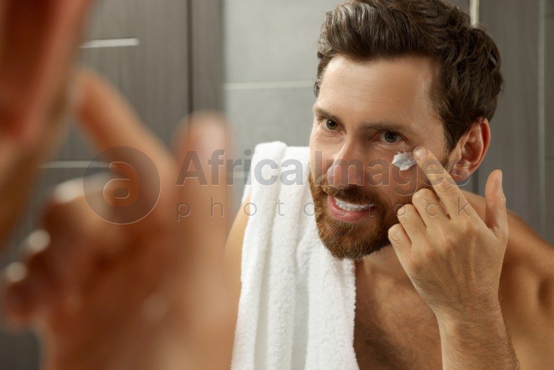 Handsome man applying cream on face in bathroom near mirror