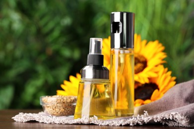 Spray bottles with cooking oil near sunflower seeds and flowers on wooden table against blurred green background