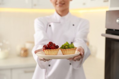 Happy professional confectioner holding plate with delicious tartlets in kitchen, closeup