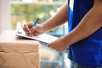 Young courier with parcels and clipboard against blurred background, closeup. Delivery service