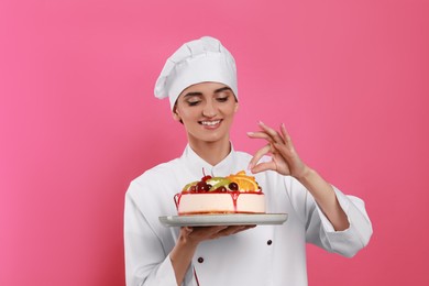 Photo of Happy professional confectioner in uniform decorating delicious cake on pink background
