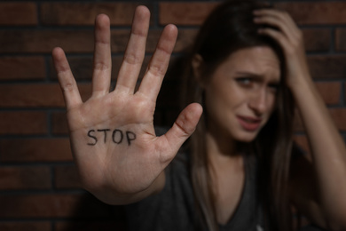 Abused young woman showing palm with word STOP near brick wall, focus on hand. Domestic violence concept