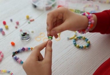 Girl making beaded jewelry at white wooden table, closeup
