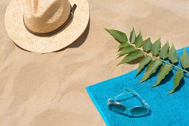 Photo of Soft blue beach towel, sunglasses, straw hat and green leaves on sand, above view