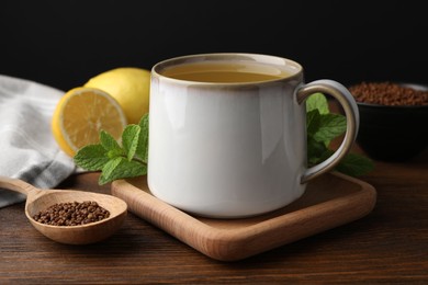Cup of aromatic buckwheat tea, spoon with granules, lemon and mint on wooden table, closeup