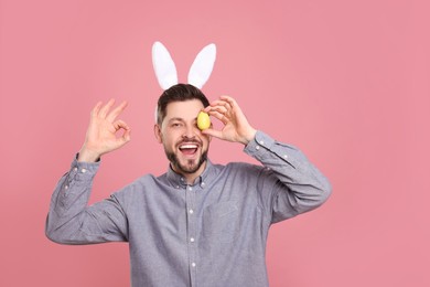 Photo of Happy man in bunny ears headband holding painted Easter egg on pink background, space for text