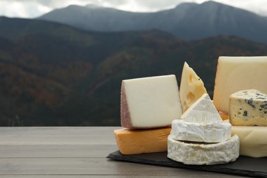 Different types of delicious cheeses on wooden table against mountain landscape. Space for text