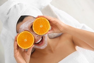 Pretty woman with rejuvenating facial mask holding sliced orange in spa salon, above view