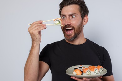 Emotional man eating tasty sushi roll and holding plate with food against light grey background, closeup