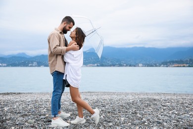 Young couple with umbrella enjoying time together under rain on beach, space for text