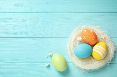Colorful Easter eggs in decorative nest and flowers on light blue wooden background, flat lay. Space for text