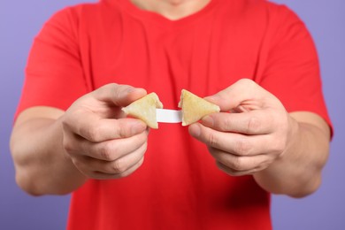 Photo of Man holding tasty fortune cookie with prediction on violet background, closeup