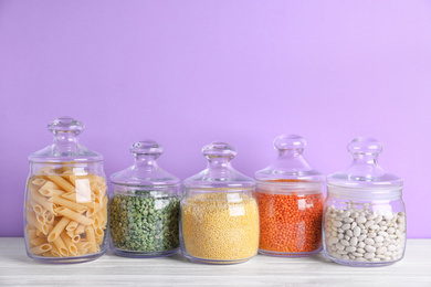 Jars with different cereals on white wooden table against violet background