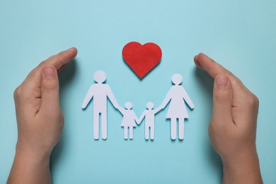 Man protecting paper family figures and red heart on light blue background, top view. Insurance concept