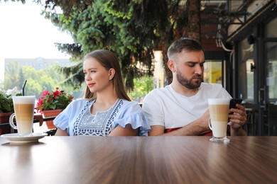 Young man with smartphone ignoring his girlfriend in outdoor cafe. Boring date