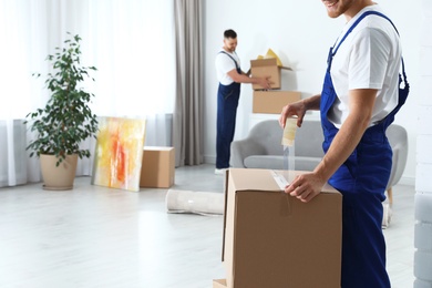 Moving service employee sealing cardboard box with adhesive tape in room, closeup