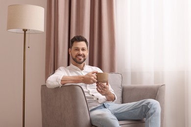 Smiling man drinking tea while resting on armchair near window with beautiful curtains at home. Space for text