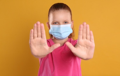 Little girl in protective mask showing stop gesture on yellow background. Prevent spreading of coronavirus