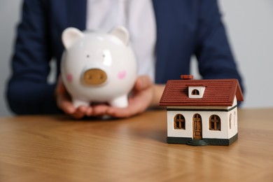 Photo of Woman holding piggy bank at wooden table, focus on little house model