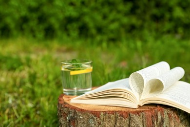 Open book near glass of water with mint and lemon on tree stump outdoors. Space for text