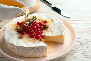 Pouring honey onto brie cheese served with almonds and red currants on white wooden table, closeup