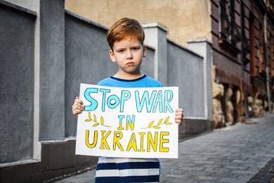 Sad boy holding poster in colors of national flag with words Stop War In Ukraine on city street