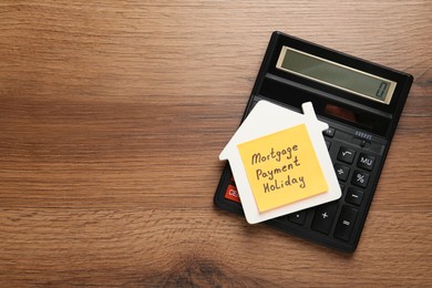 Photo of Paper note with words Mortgage Payment Holiday, house model and calculator on wooden table, top view. Space for text
