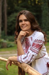 Beautiful woman wearing embroidered shirt near wooden railing in countryside. Ukrainian national clothes
