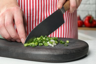 Woman cutting green spring onion on black wooden board at white table, closeup