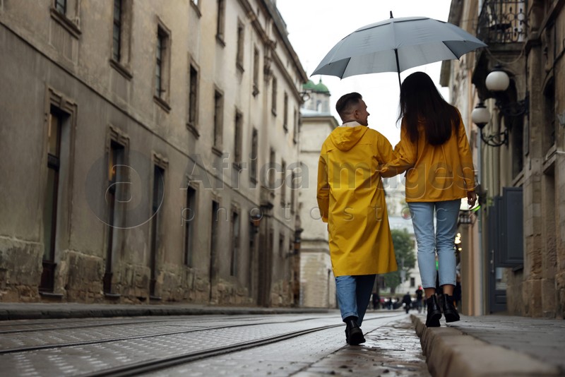 Photo of Lovely young couple with umbrella walking under rain on city street, back view